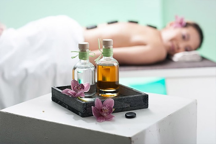 Tray mit zwei Fläschen Massageöl und Orchideeblüten, im Hintergrund liegt eine thailändische Frau auf dem Bauch und bekommt eine Hot Stone-Massage